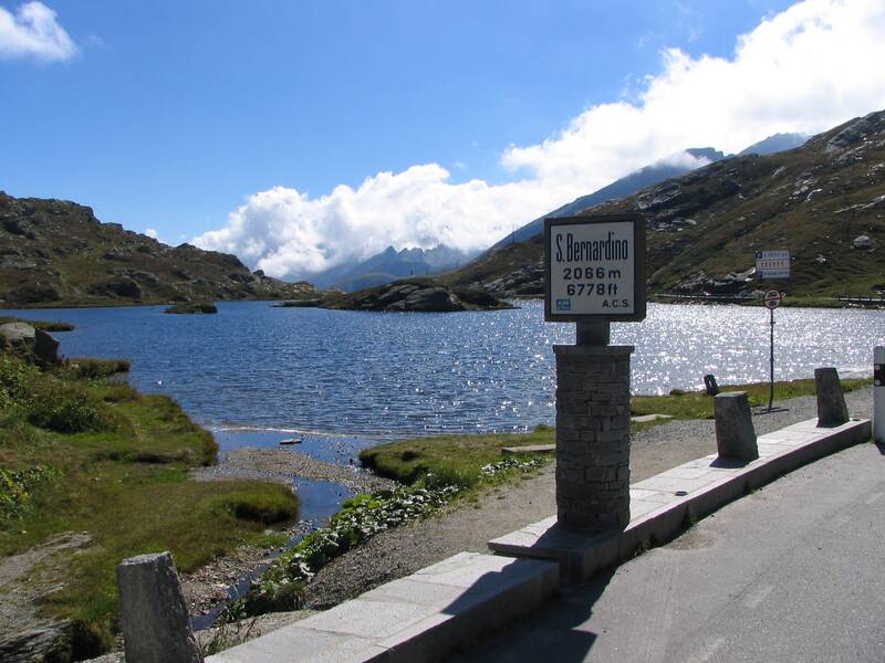 Ein klarer See liegt in einer bergigen Landschaft unter blauem Himmel, daneben steht ein Schild mit der Aufschrift "S. Bernardino 2066 m 6778 ft".