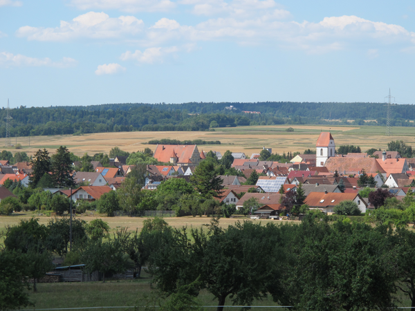 Eine malerische Dorfansicht mit roten Dächern und einer Kirche, umgeben von grünen Feldern und Bäumen unter einem blauen Himmel mit wenigen Wolken.