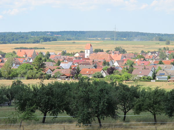 Eine malerische Dorflandschaft mit zahlreichen rotgedeckten Häusern und einer Kirche, umgeben von grünen Feldern und Bäumen.