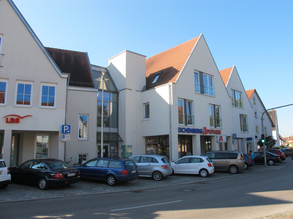 Eine Straße mit einreihig geparkten Autos vor einem modernen Gebäude, in dem eine Apotheke und ein weiteres Geschäft untergebracht sind.