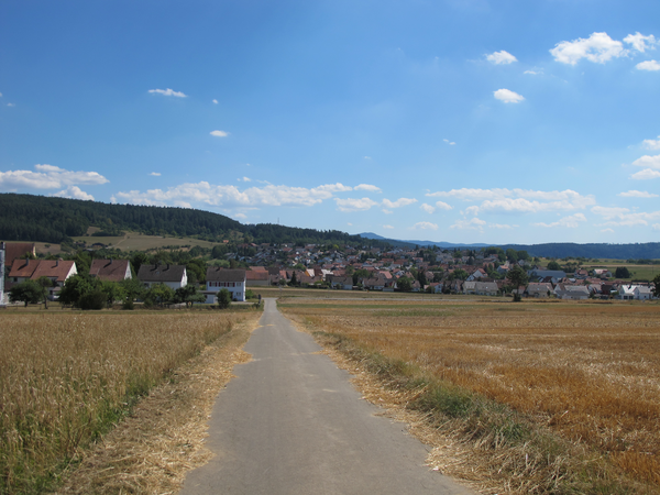 Ein asphaltierter Weg führt durch weite Felder auf ein Dorf mit roten Dächern zu, umgeben von Wäldern und Hügeln unter einem blauen Himmel.