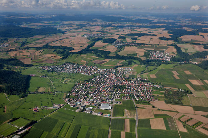 Luftaufnahme einer weitläufigen Landschaft mit einem Dorf in der Mitte, umgeben von Feldern und Wäldern.