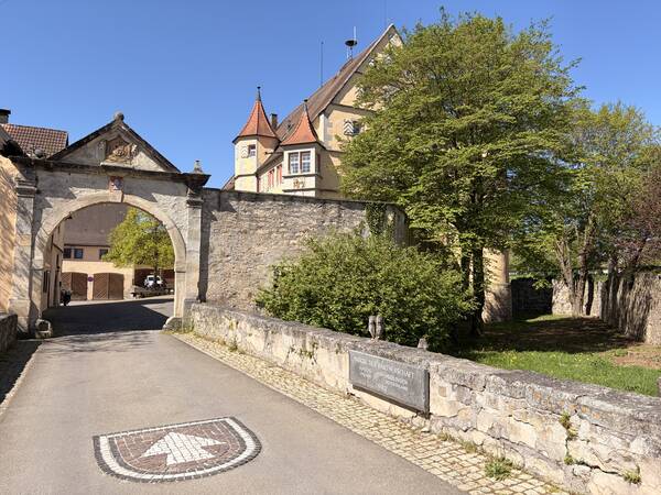 Ein steinerner Torbogen führt zu einem Innenhof mit historisch anmutenden Gebäuden und einer Baumreihe an der Seite unter einem klaren blauen Himmel.