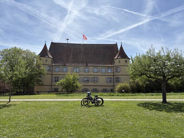Vor einem historischen Gebäude mit zwei Türmen und einer österreichischen Flagge auf dem Dach stehen zwei Fahrräder auf einer grünen Wiese, umgeben von Bäumen.