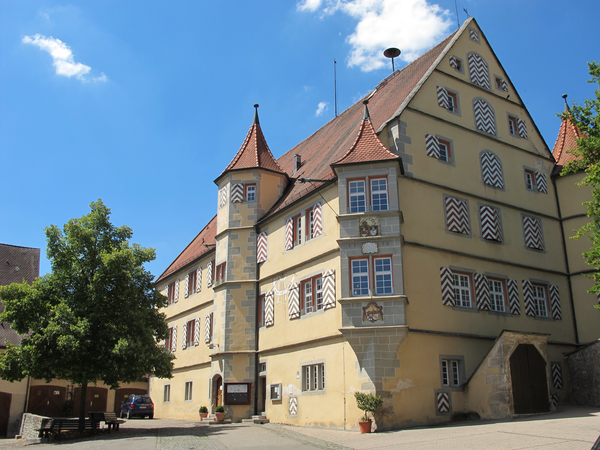 Historisches Gebäude im Fachwerkstil mit verzierten Fensterläden und einem kleinen Turm, bei blauem Himmel und Sonnenschein.