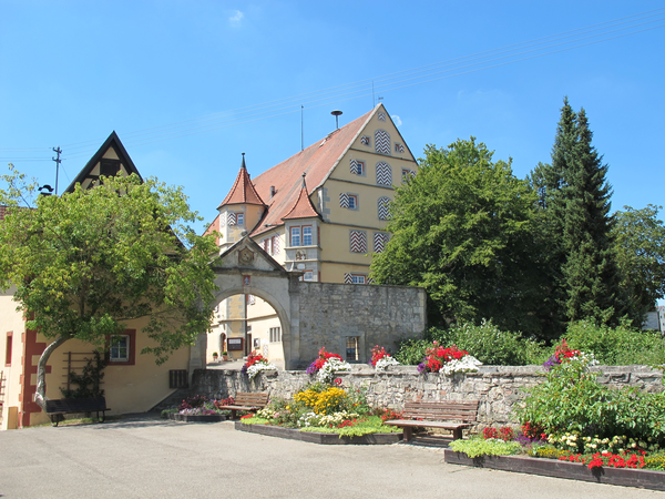 Ein historischer Torbogen führt durch eine Steinmauer zu einem Gebäude mit bunt gemusterten Fenstern, umrahmt von blühenden Blumen und Bäumen.