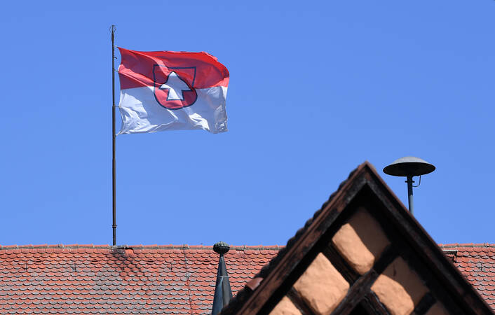 Eine rot-weiße Flagge mit einem Wappenmotiv weht auf einem Gebäude mit ziegelrotem Dach vor strahlend blauem Himmel.