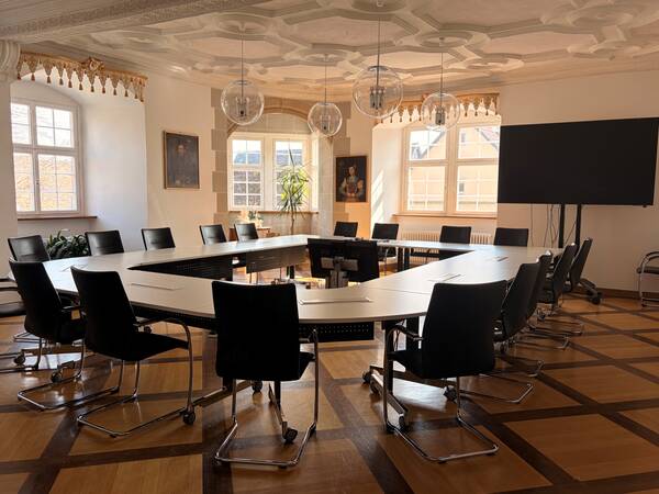 Ein Konferenzraum mit einem großen ovalen Tisch, umgeben von schwarzen Stühlen, mit einem großen Bildschirm an der Wand und mehreren Fenstern, die Tageslicht hereinlassen.