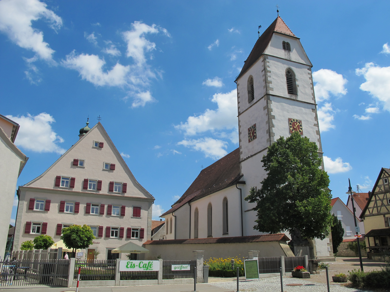 Ein kleines Dorf mit einer Kirche im Zentrum, flankiert von traditionellen Gebäuden und einem Eis-Café im Vordergrund, unter einem sonnigen, blauen Himmel.